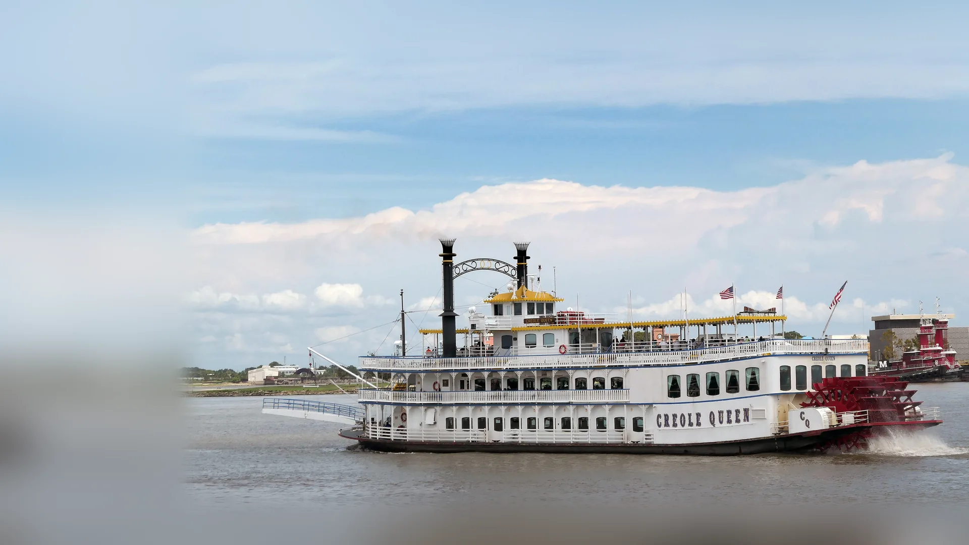 Den hvide hjuldamper Creole Queen på Mississippi-floden, med to sorte skorstene, et øvre dæk med gule kanter og et rødt hjulet hjul i agterstavnen, under en blå himmel med en rød slæbebåd bagved