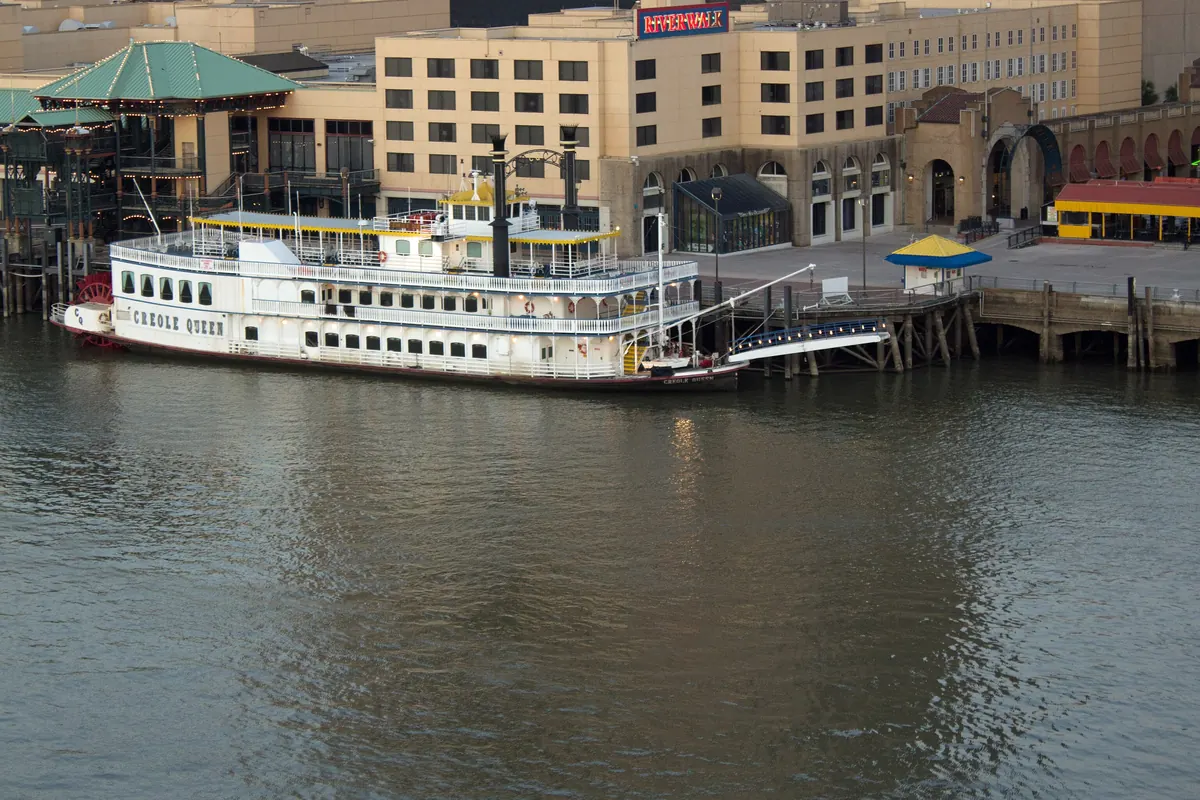 La Creole Queen ormeggiata al molo del Riverwalk a New Orleans, sotto una fila di edifici alberghieri e commerciali, con l'acqua bruna del fiume in primo piano