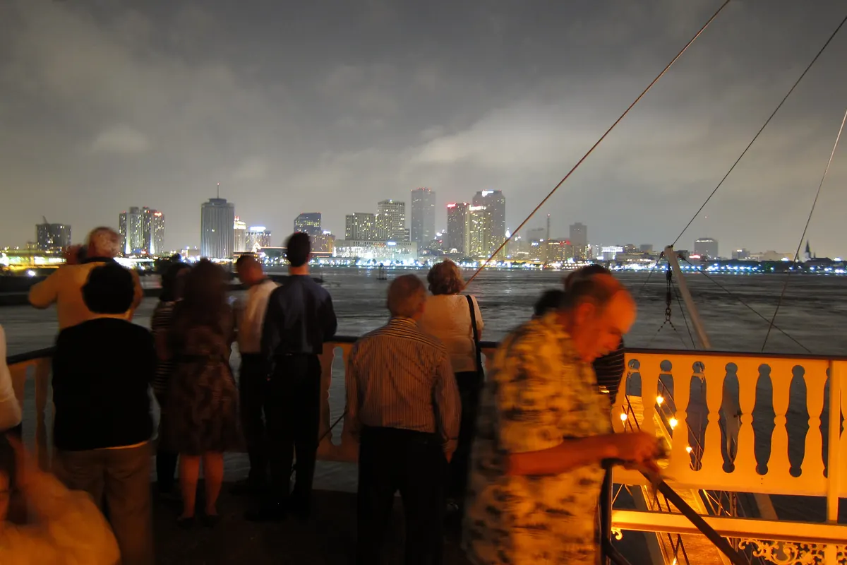 Passeggeri alla ringhiera del ponte di un battello fluviale di notte che guardano oltre il Mississippi lo skyline illuminato di New Orleans