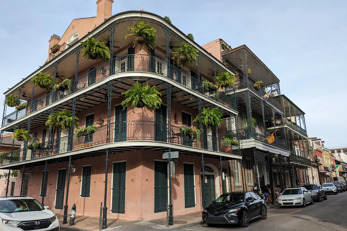 Un edificio d'angolo rosa di tre piani nel French Quarter con balconi curvi in ghisa adornati di felci, e auto parcheggiate sulla strada sottostante